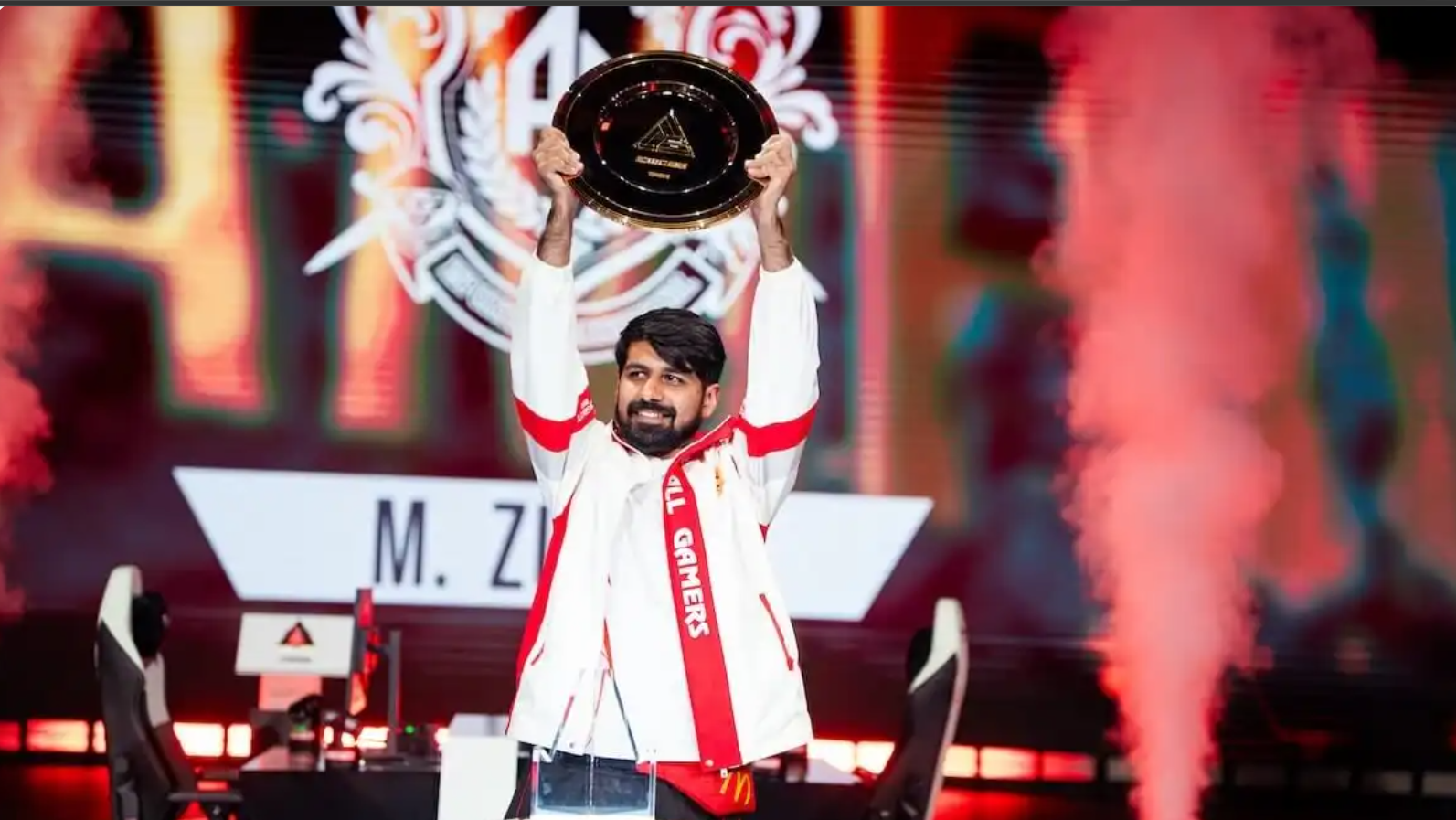 M. Zubair: New Tekken Champion Emerged Professional esports player M. Zubair stands center stage smiling as he holds a large black and gold championship trophy disc high above his head. He is wearing a white and red All Gamers (AG) team jacket. The background features a large LED screen displaying his name "M. ZUBAIR" along with his team's logo, framed by dramatic pillars of orange and red stage smoke.